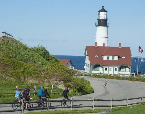 Four people ride bicycles along a paved path near a white lighthouse with a red roof by the ocean. The sky is clear and blue, and an American flag flies nearby. Green shrubs and wooden fences border the path.