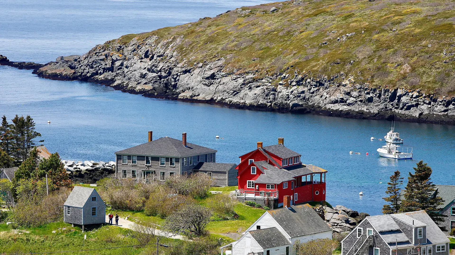 Colorful houses near a rocky shoreline overlook a calm bay with a boat anchored in the water; green hills and sparse trees surround the peaceful coastal village.