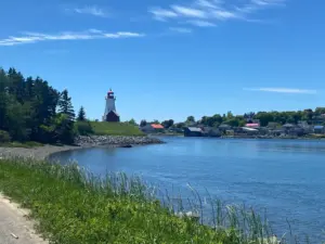 A scenic view of a coastal village with a lighthouse on a grassy hill, surrounded by trees and houses. Calm blue water is in the foreground, under a bright blue sky with wispy clouds.