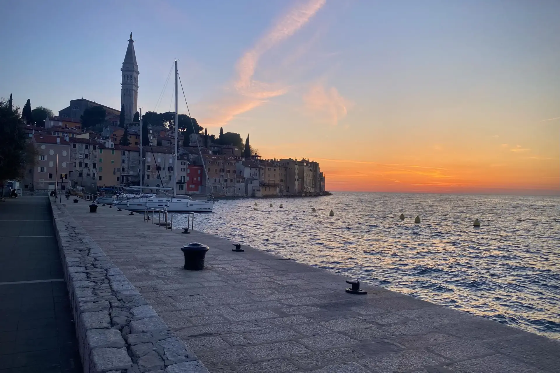 Stone waterfront with boats docked beside colorful buildings and a tall church tower at sunset. The sky glows orange and pink, and the calm sea reflects the fading light.
