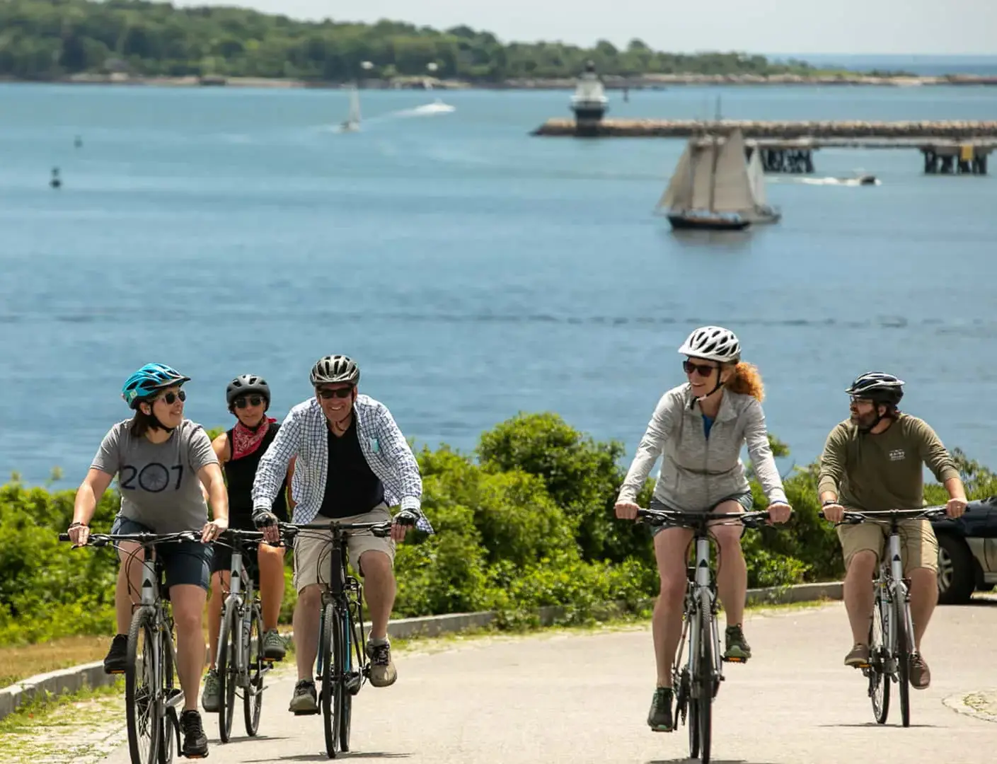Five people ride bicycles on a paved path by the water, wearing helmets and casual clothes. In the background, sailboats and a pier are visible on a sunny day.