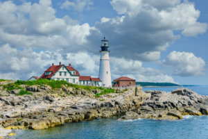 A white lighthouse with a black top stands on a rocky shoreline next to red-roofed buildings, under a blue sky filled with puffy clouds. The ocean is calm in the foreground.