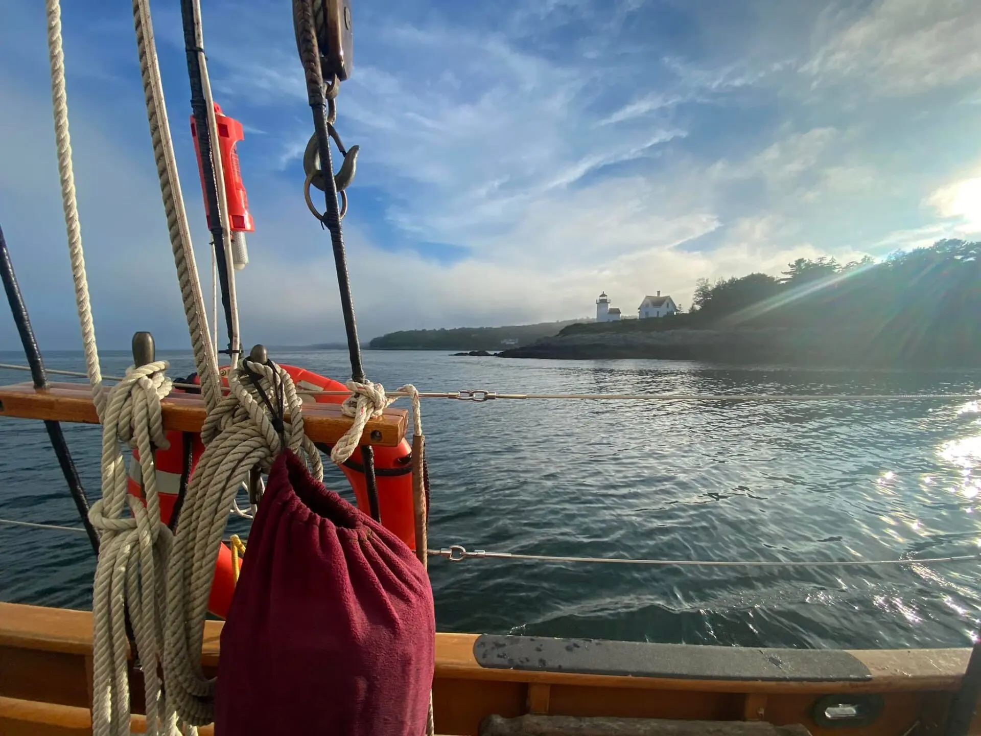 View from a sailboat with ropes and a red bag in the foreground, looking toward a distant white lighthouse on a rocky, tree-lined shore under a blue sky with scattered clouds.