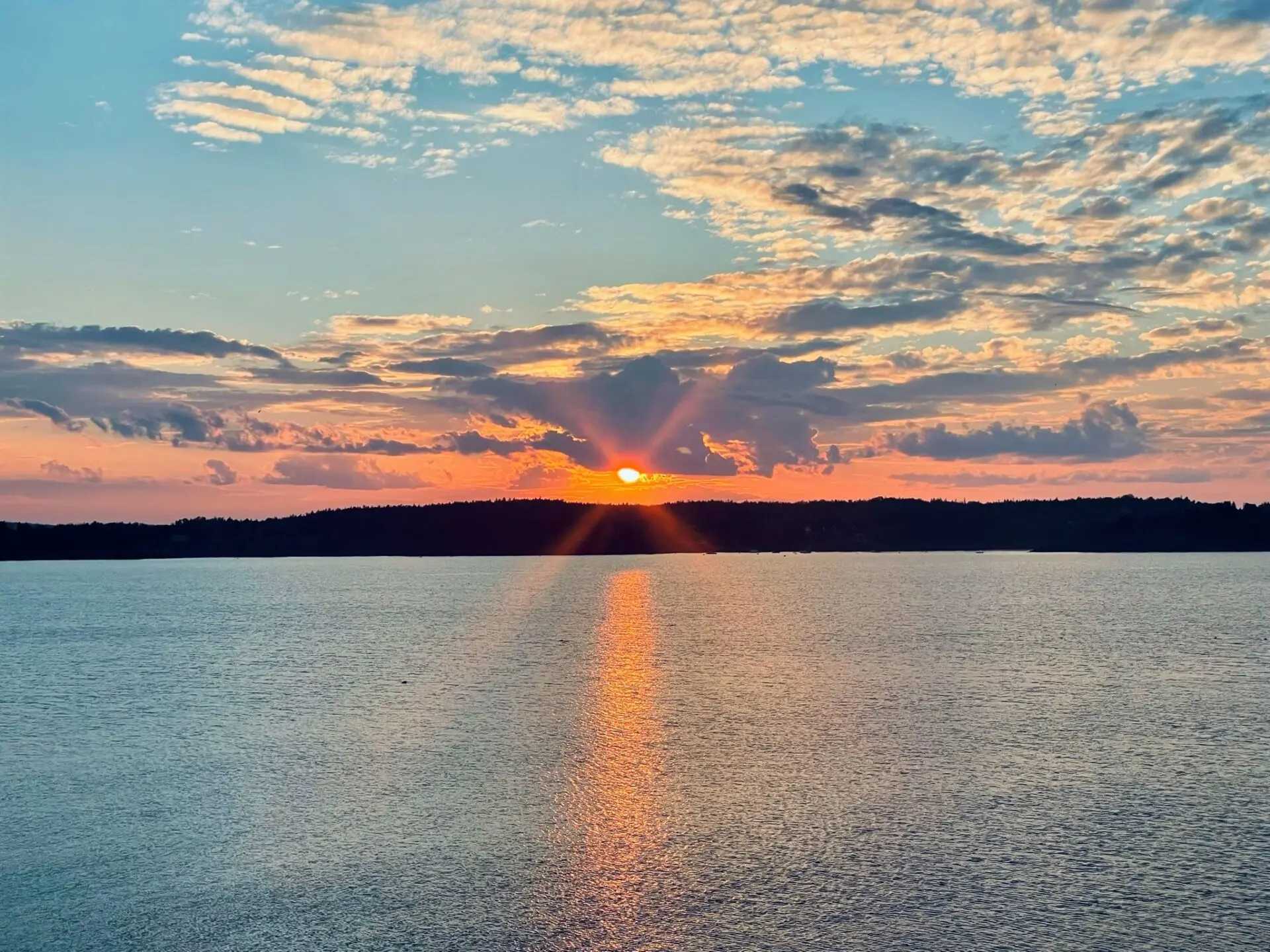 A vibrant sunset over a calm body of water, with the sun partially hidden behind a distant treeline. Orange and pink rays reflect on the water, while scattered clouds color the sky in shades of blue, orange, and yellow.