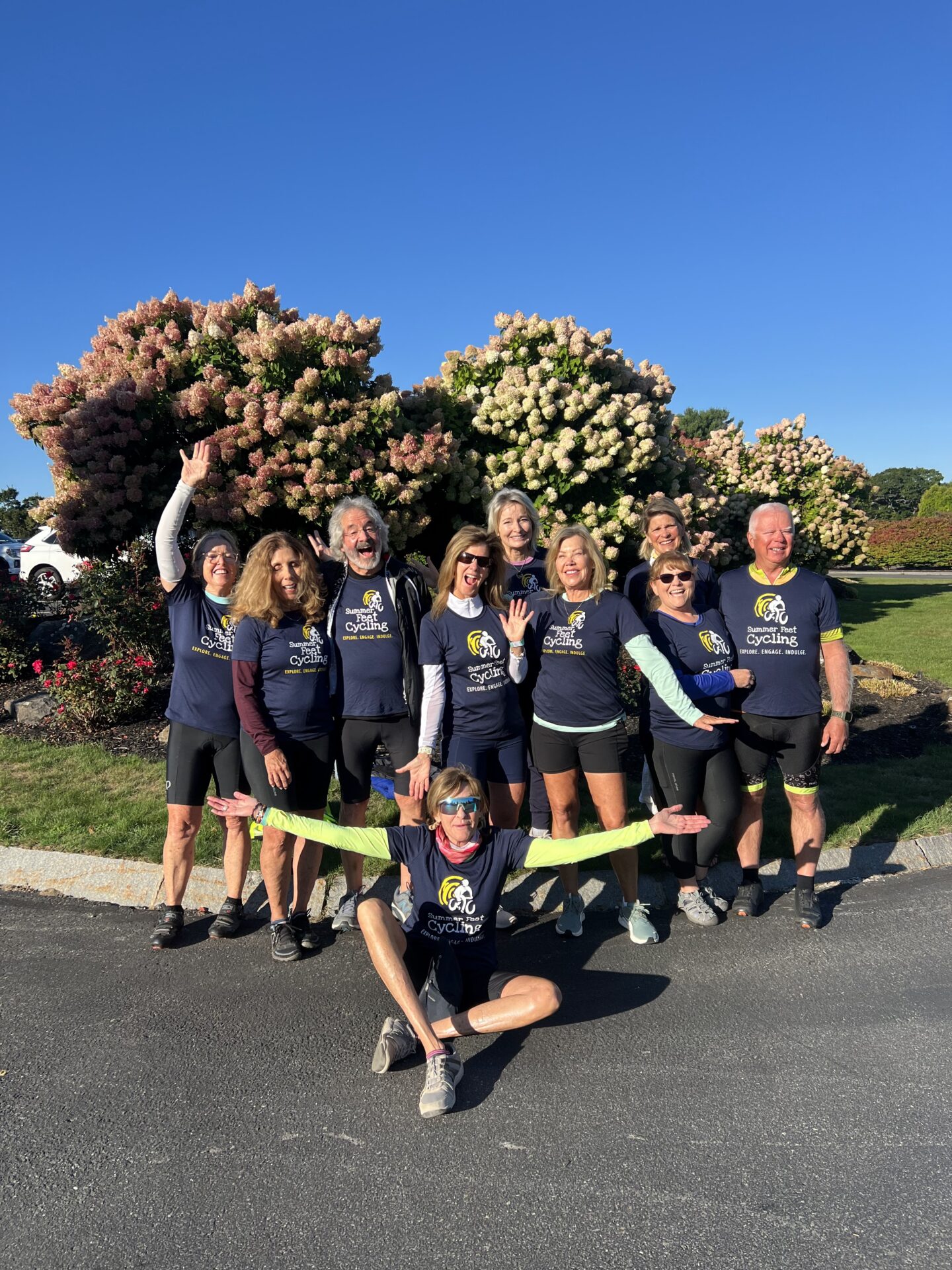 A group of ten adults in matching cycling shirts pose and smile outdoors on a sunny day, standing and sitting in front of blooming bushes with pink and white flowers.