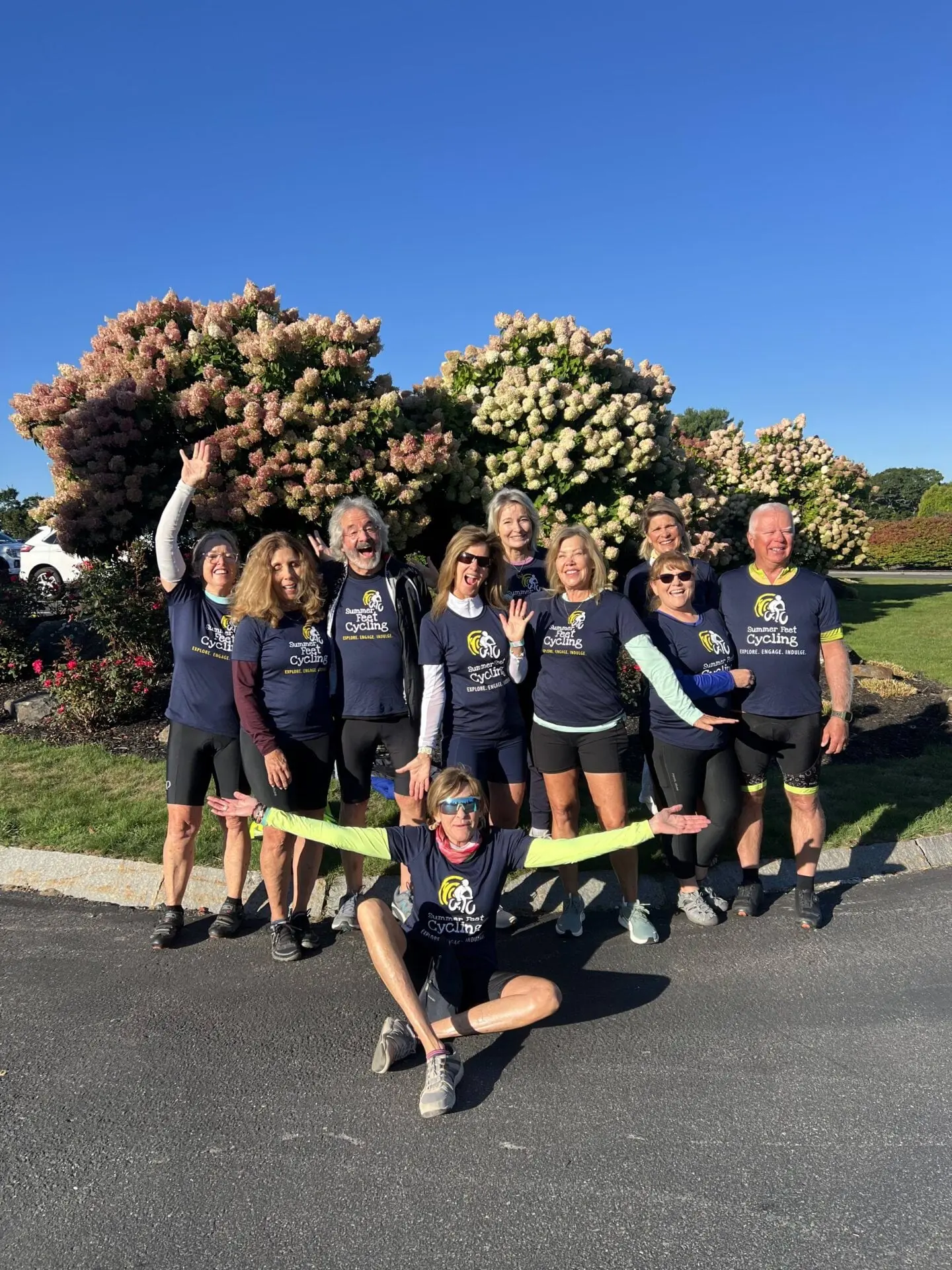 A group of ten smiling adults in matching cycling shirts pose together outside on a sunny day, celebrating their adventure with Bike Tour Gift Certificates. Blooming bushes and a clear blue sky complete the cheerful scene.