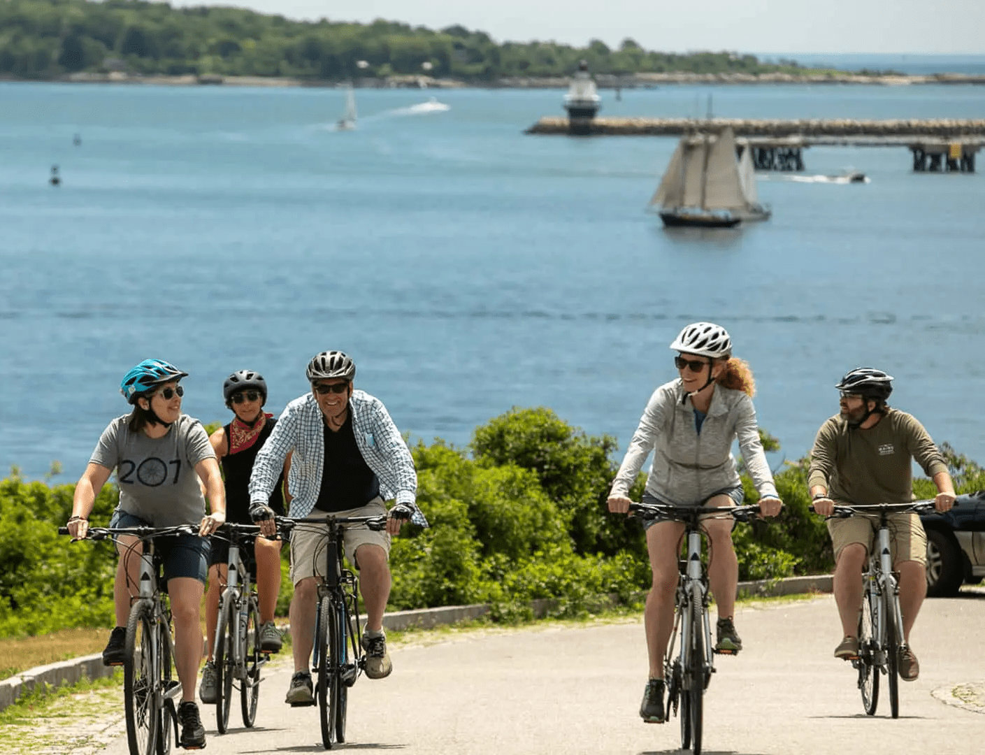 Five people ride bicycles on a paved path near the water, smiling and talking. Green bushes, a sailboat, and a pier are visible in the background under a clear blue sky.