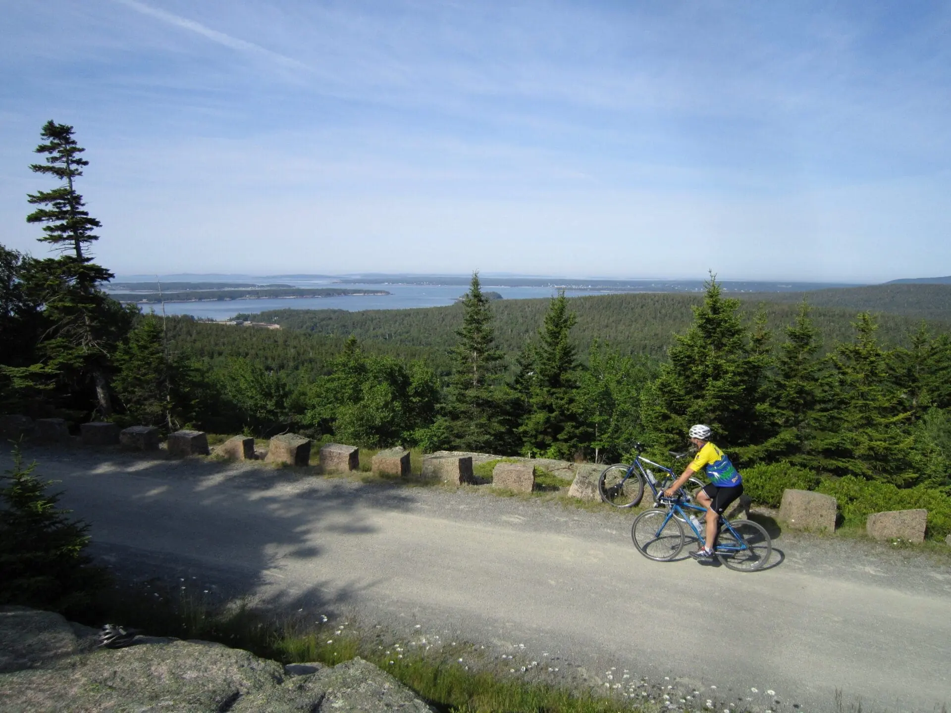 A cyclist wearing a helmet rides along a gravel path bordered by trees, with a scenic view of a forest and water in the distance under a clear blue sky.