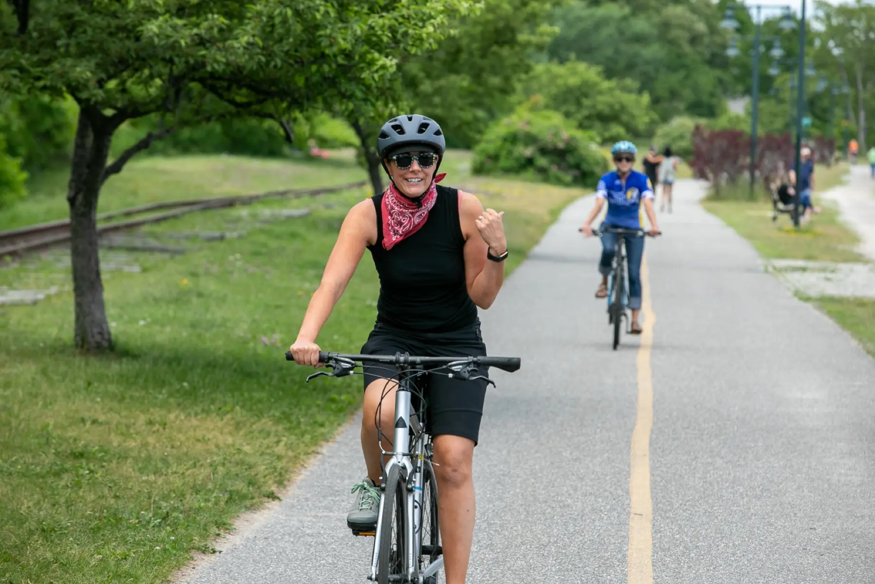 A smiling woman wearing a helmet, sunglasses, and a red bandana rides a bike on a paved path, while other cyclists follow behind her on a sunny day surrounded by greenery.